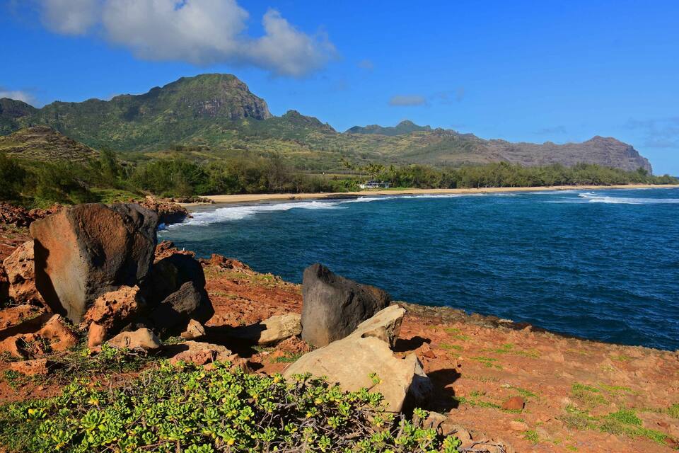 A coastal landscape with rocky shores, a sandy beach, and lush green mountains under a clear blue sky.