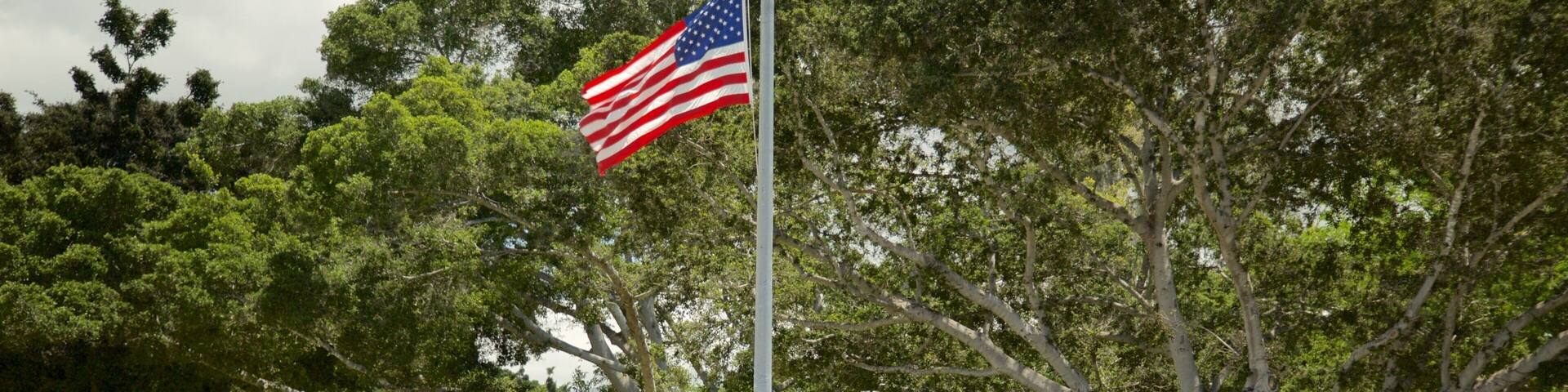 Un asta con la bandera estadounidense, un cartel que dice "USS OKLAHOMA BB-37" y una cerca blanca de piquetes.