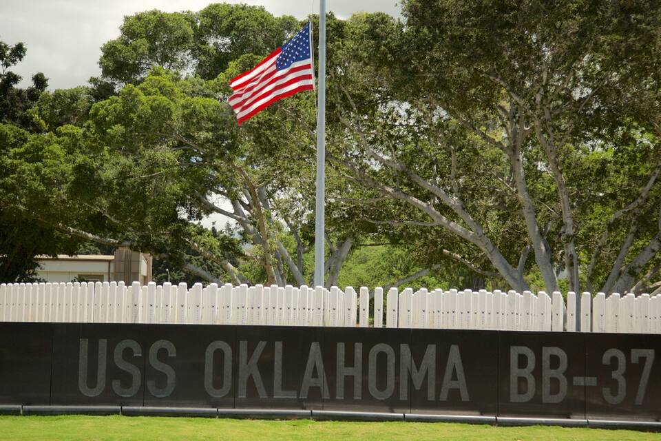 A flagpole with an American flag, a sign reading "USS OKLAHOMA BB-37", and a white picket fence.