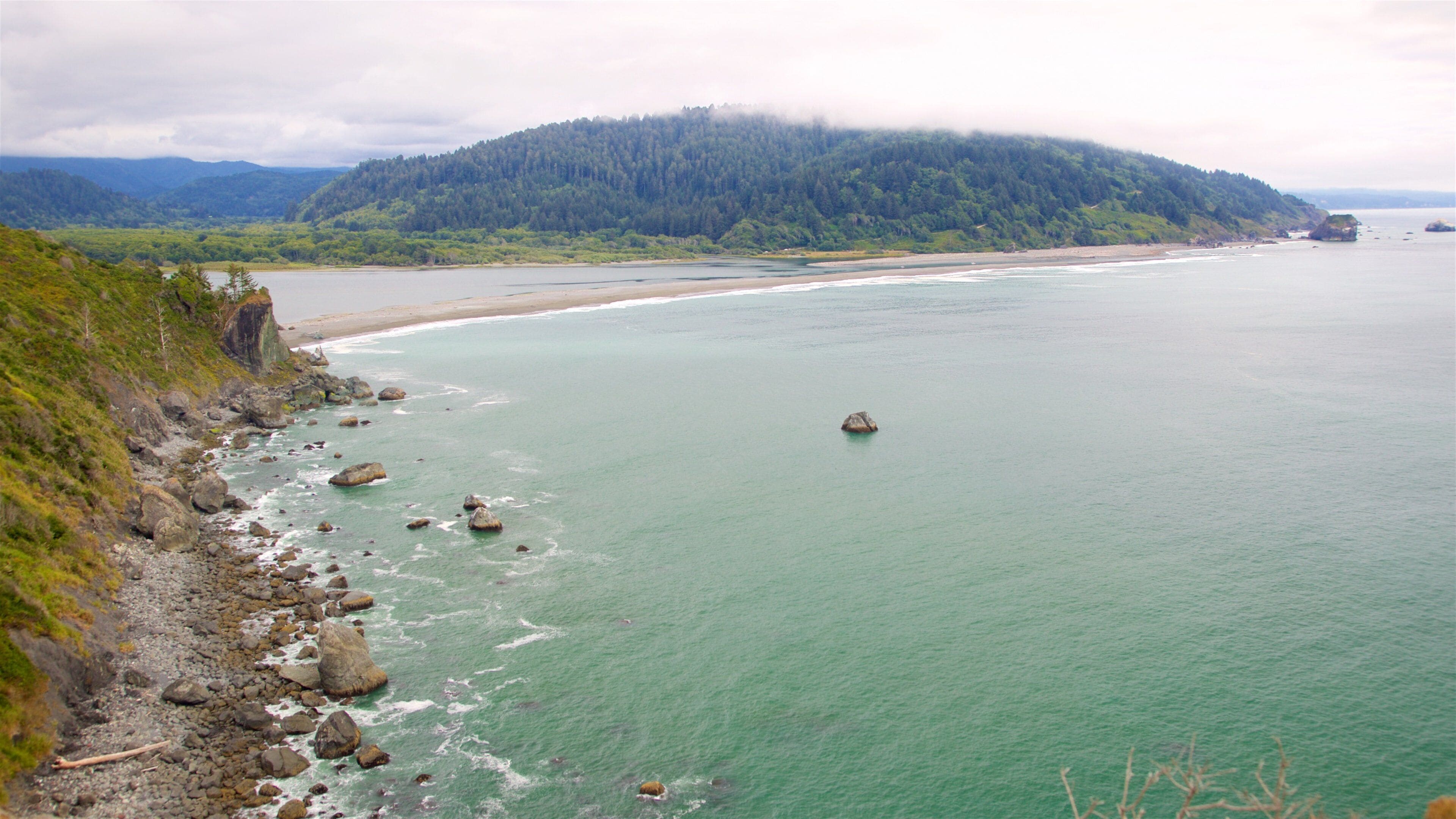 A coastal landscape with a rocky shoreline, calm turquoise waters, and a forested hill in the background.