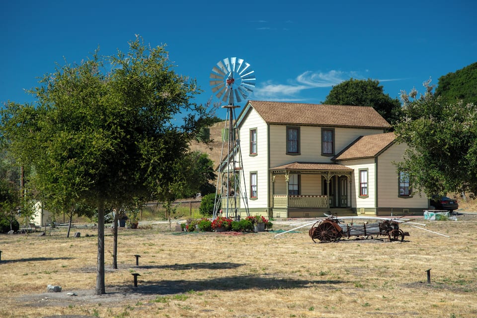 Une maison historique avec un moulin à vent, entourée d’arbres et bordée d’un champ aride.