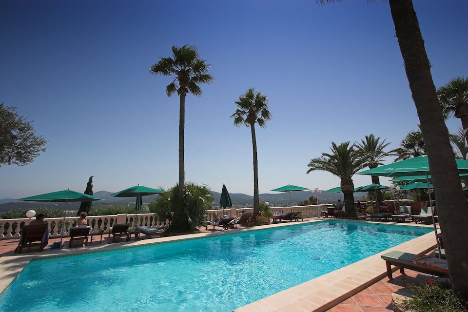 A pool with lounge chairs and umbrellas, surrounded by palm trees and a clear sky.