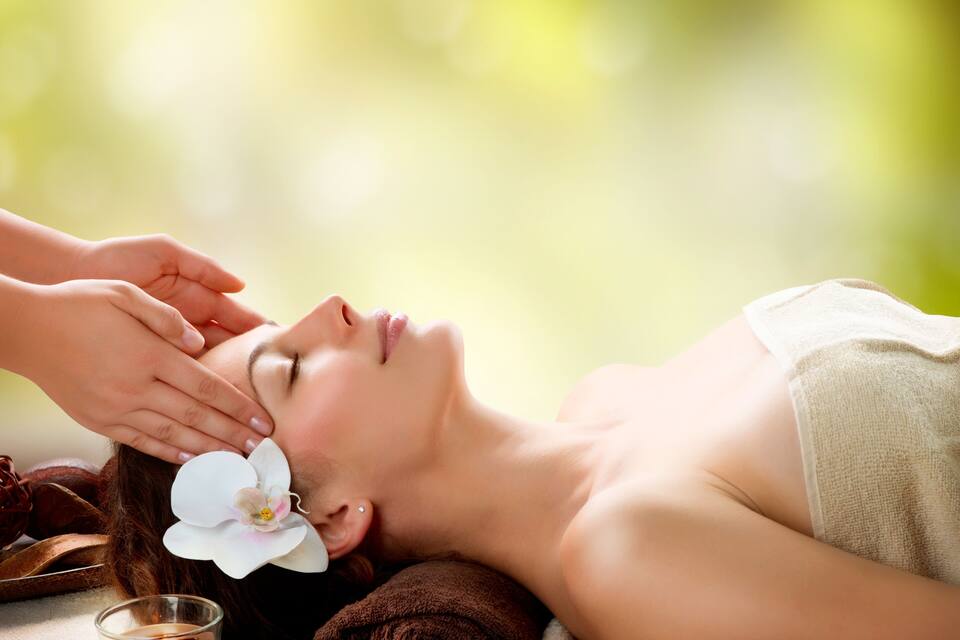 Une personne qui reçoit un soin du visage, avec une fleur dans les cheveux, entourée de pierres et d’une bougie.