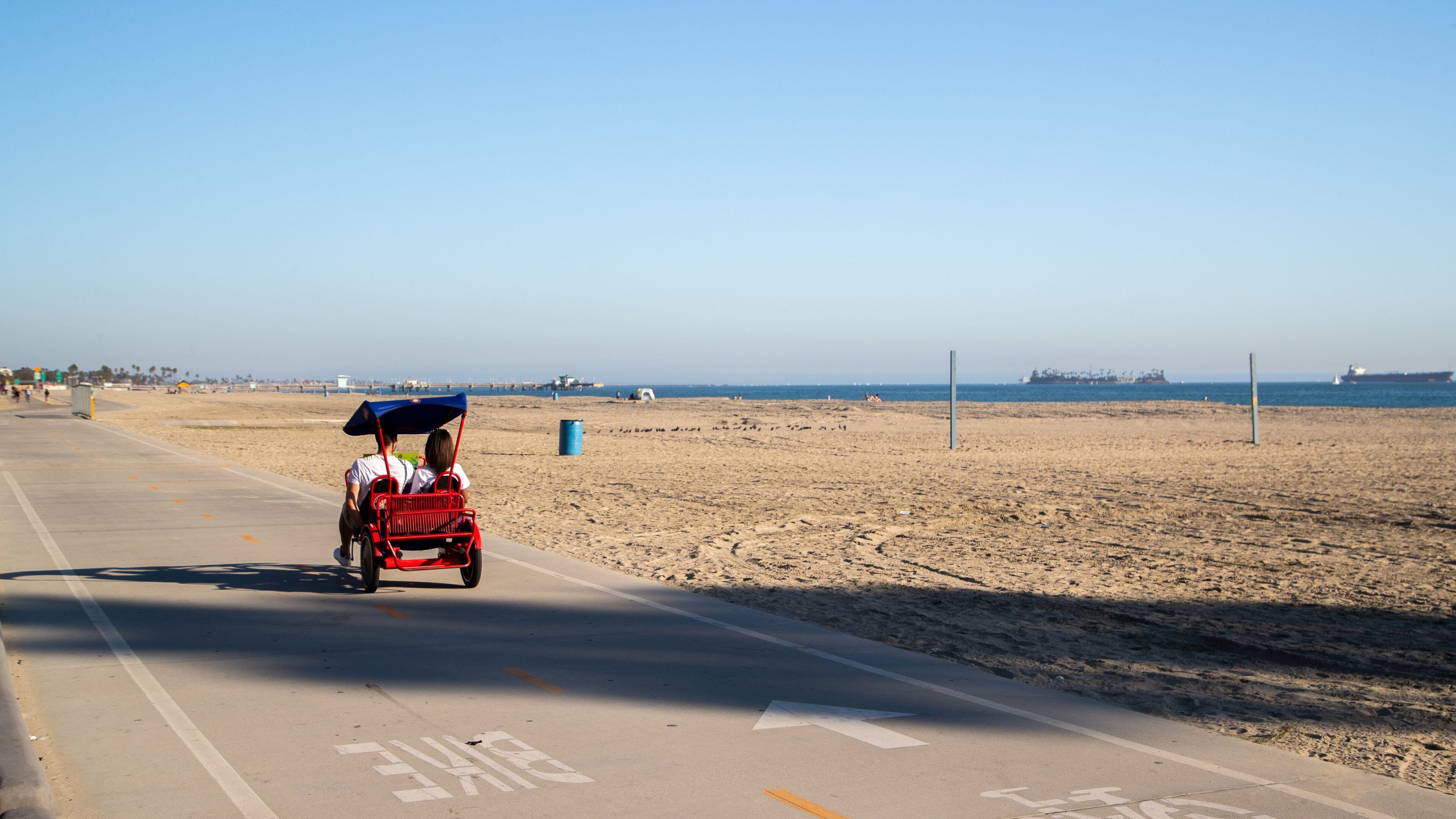 Una carretera frente a la playa con un carrito rojo, cielos despejados y un barco a lo lejos.