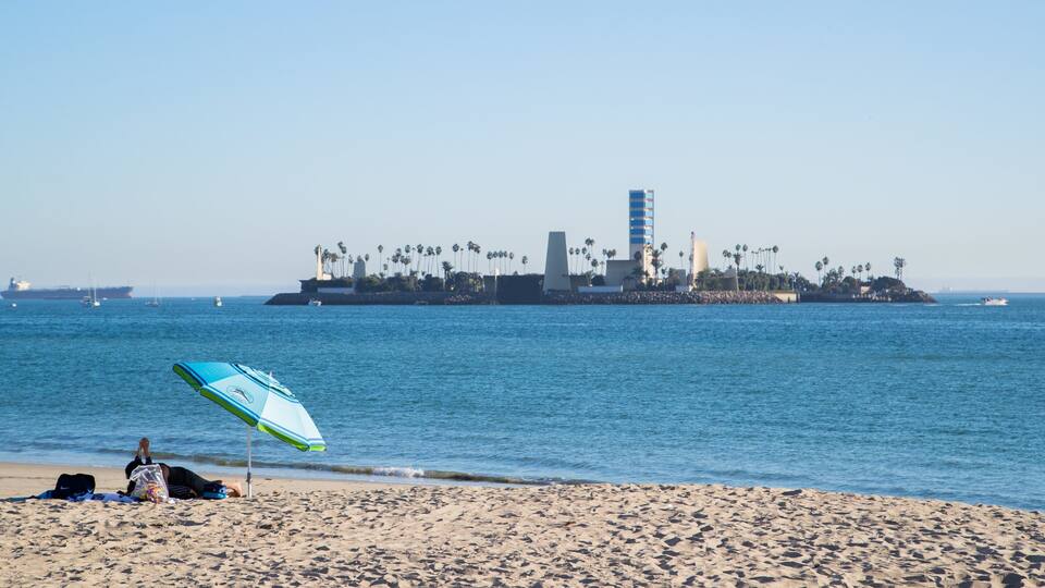 Una playa con una persona descansando bajo una sombrilla, una orilla arenosa y una isla distante con palmeras y edificios.