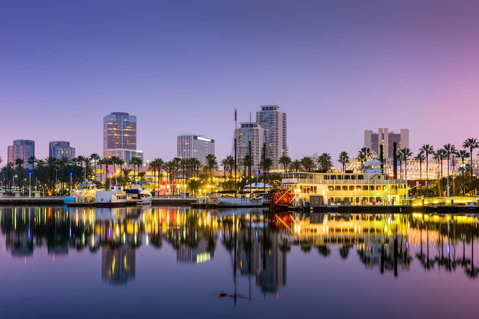 A city skyline at dusk with illuminated buildings reflected in calm waters, a boat docked near the shore, and palm trees lining the waterfront.