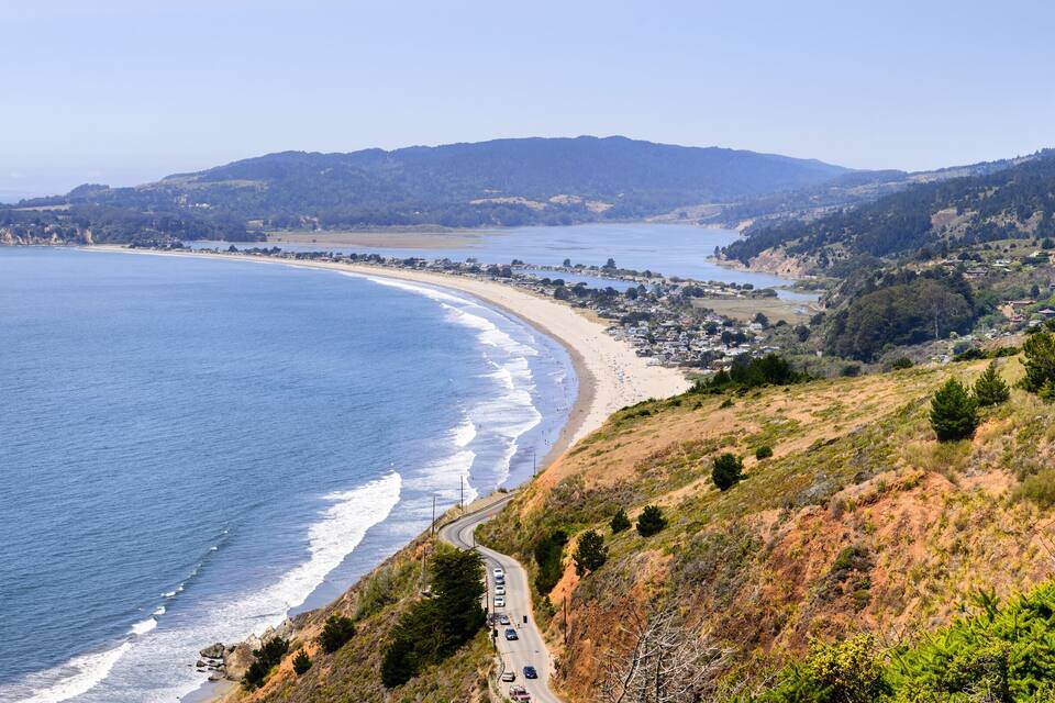 A coastal landscape with a curving road, beach, and hills.