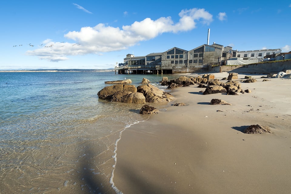 A beach with large rocks, clear water, and a building with a lighthouse in the background.