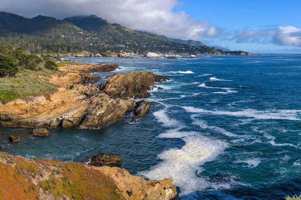 A coastal landscape with rocky cliffs, ocean waves, and a mountainous backdrop.