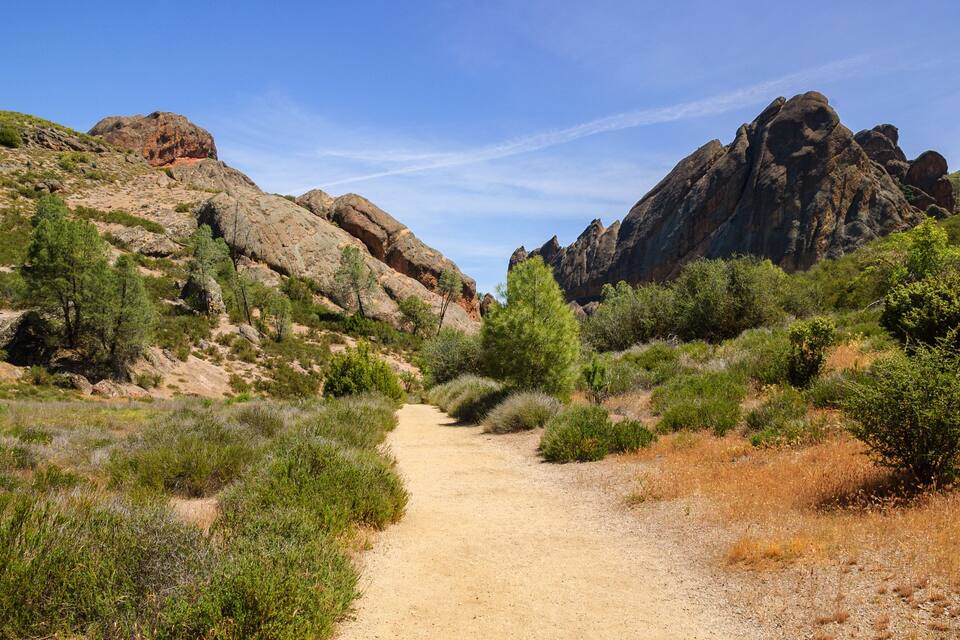 A dirt path winding through a rocky landscape with sparse vegetation and towering mountains.