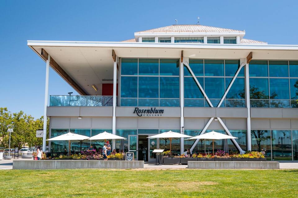 A modern building with a large glass facade, a sign reading "Rosenblum Cellars," and outdoor seating with umbrellas.
