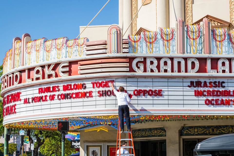 A vintage Grand Lake theater with colorful marquee lights and a sign reading "Families Belong, All People of Conscience Should Oppose."