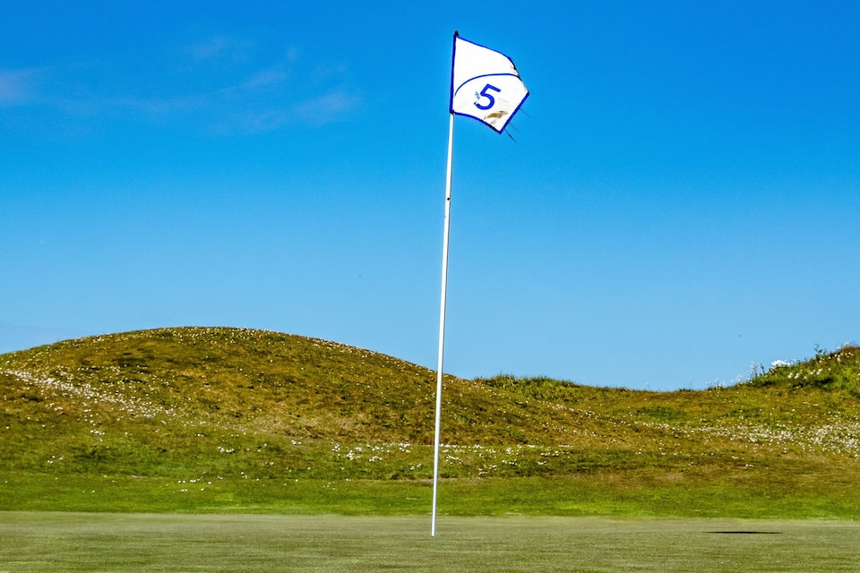 A golf course with a flag marked '5' on a flagstick, surrounded by green hills under a clear blue sky.