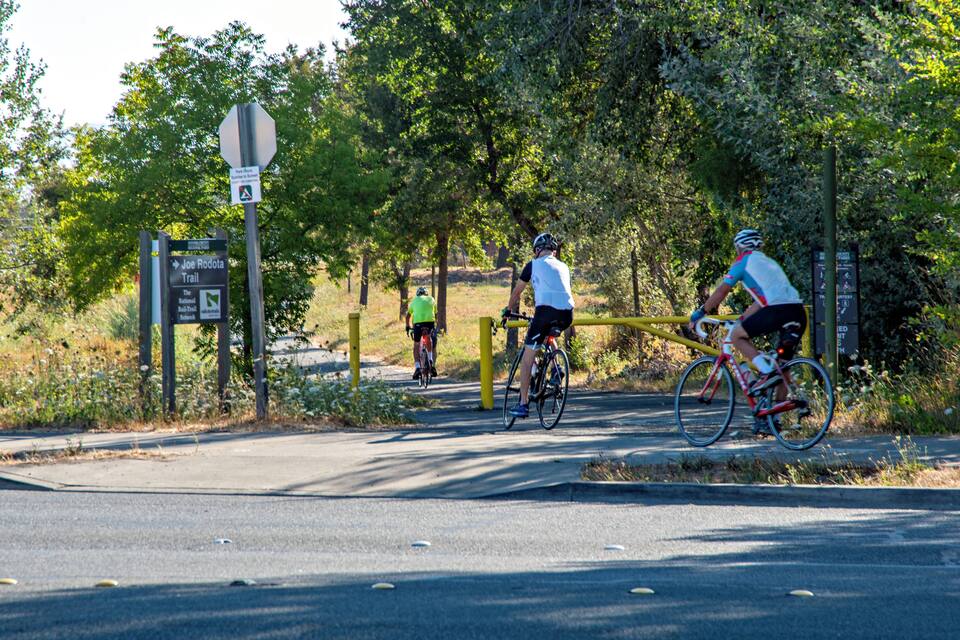 Un sendero pavimentado con ciclistas, rodeado de árboles y un cartel que indica el Joe Roddale Trail.