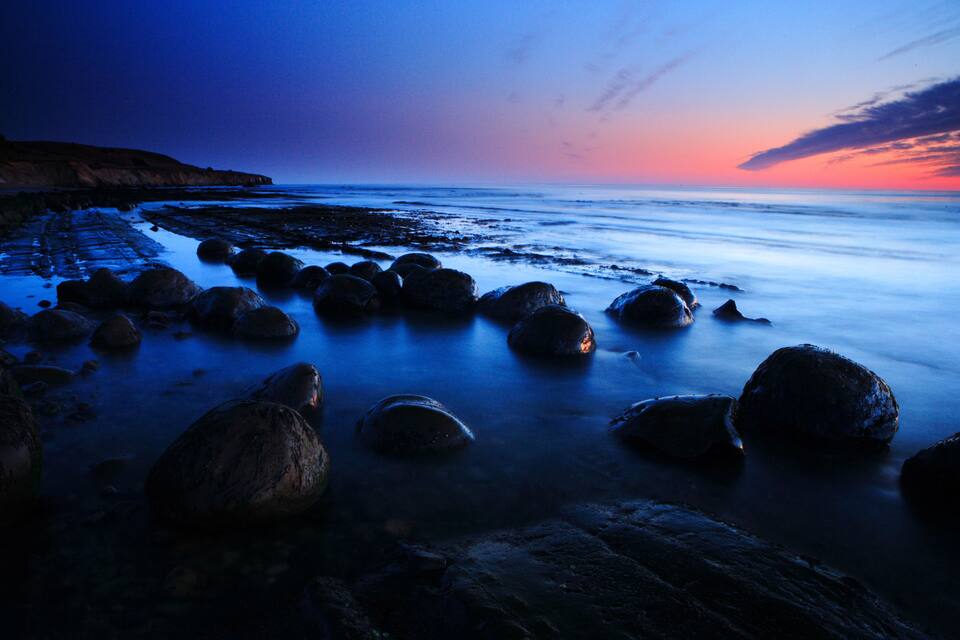 Linha de costa rochosa ao entardecer, com mar calmo e céu avermelhado pelo pôr do sol.