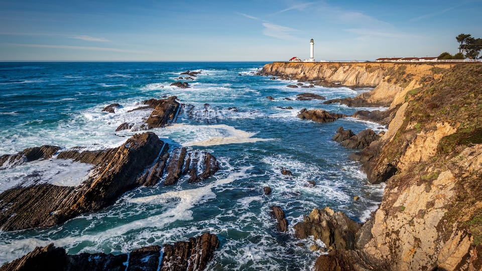 A coastal landscape with rocky cliffs, waves crashing against the shore, and a lighthouse in the distance.
