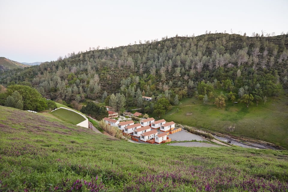 Una colina con un grupo de casas blancas de tejados rojos, rodeadas de campos verdes y una loma arbolada.