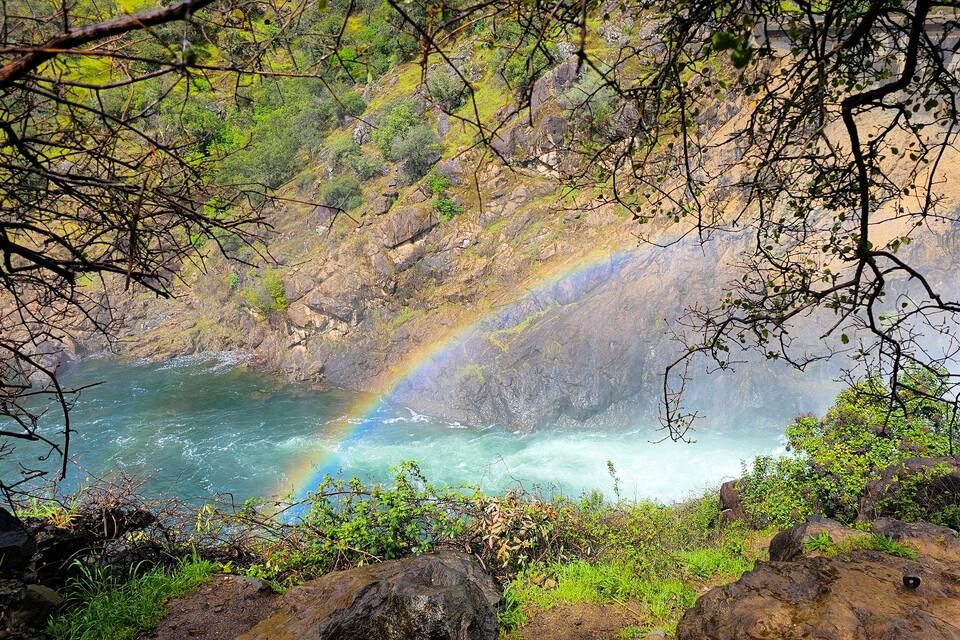 A river with a rainbow, surrounded by rocky terrain and greenery.