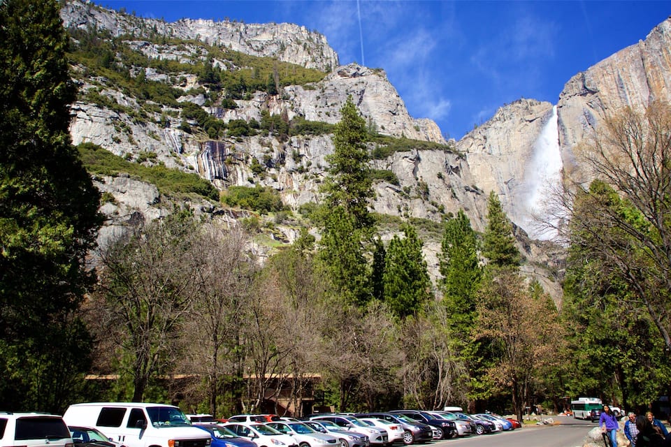 A parking lot with cars, a mountain with a waterfall, and trees.