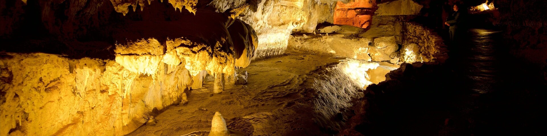 Caverna com estalactites e estalagmites, uma fogueira e uma pessoa perto da entrada.