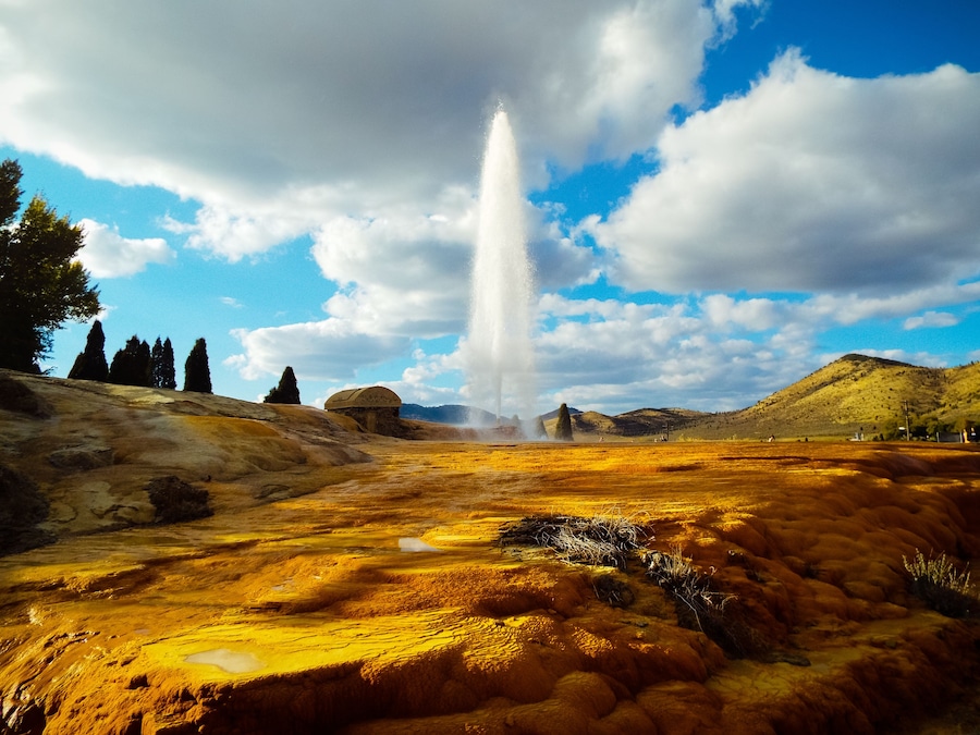 Un geyser in eruzione in una zona geotermica, con cielo sereno e colline lontane.