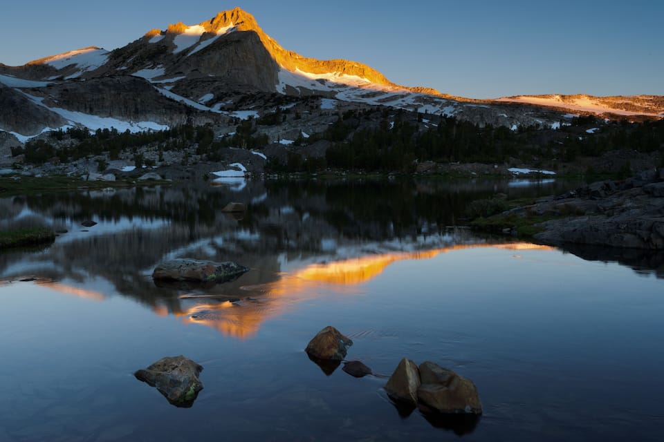 A mountain with snow patches and a calm lake with rocks in the foreground.