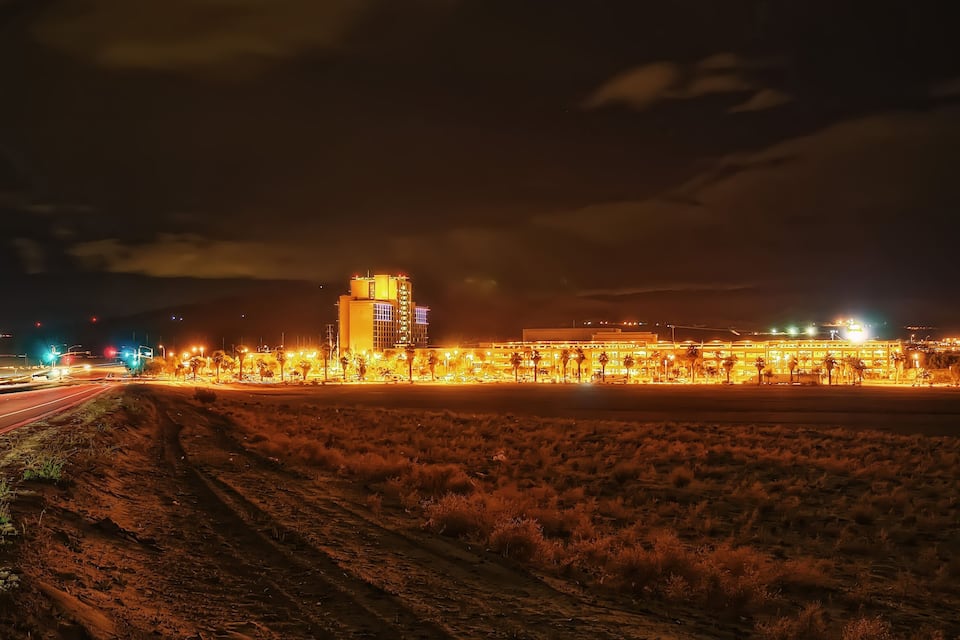 A desert landscape with a brightly lit building complex and a road with vehicles.