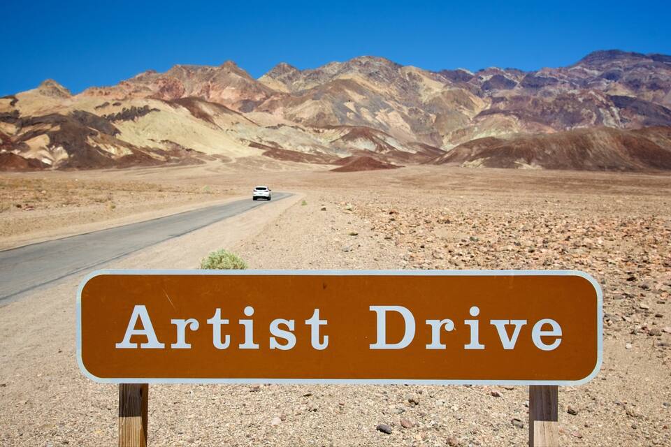 Una carretera con un cartel que dice 'Artist Drive' en un paisaje desértico con montañas al fondo.