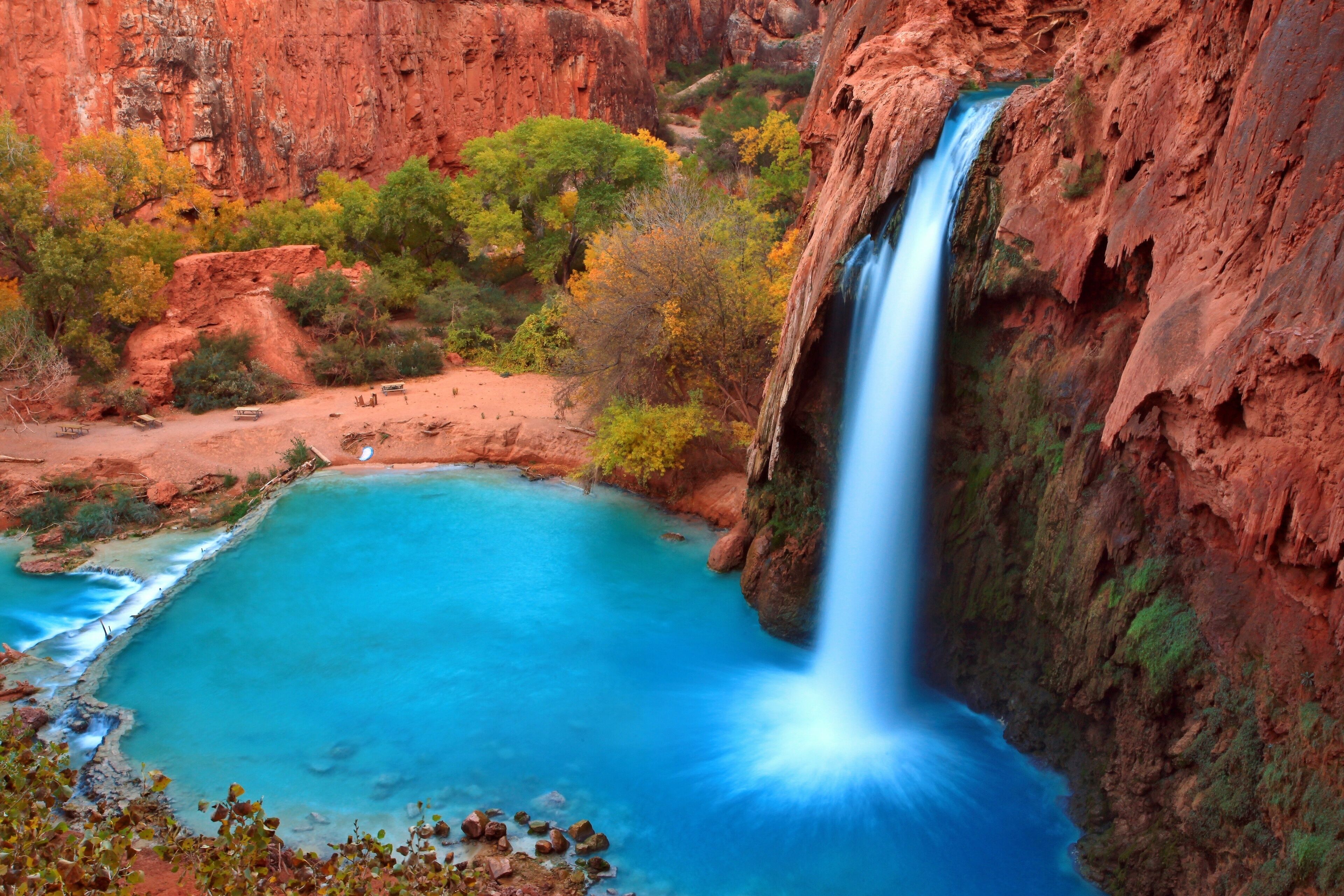 A waterfall cascading into a turquoise pool surrounded by red rock cliffs and autumn-colored trees.