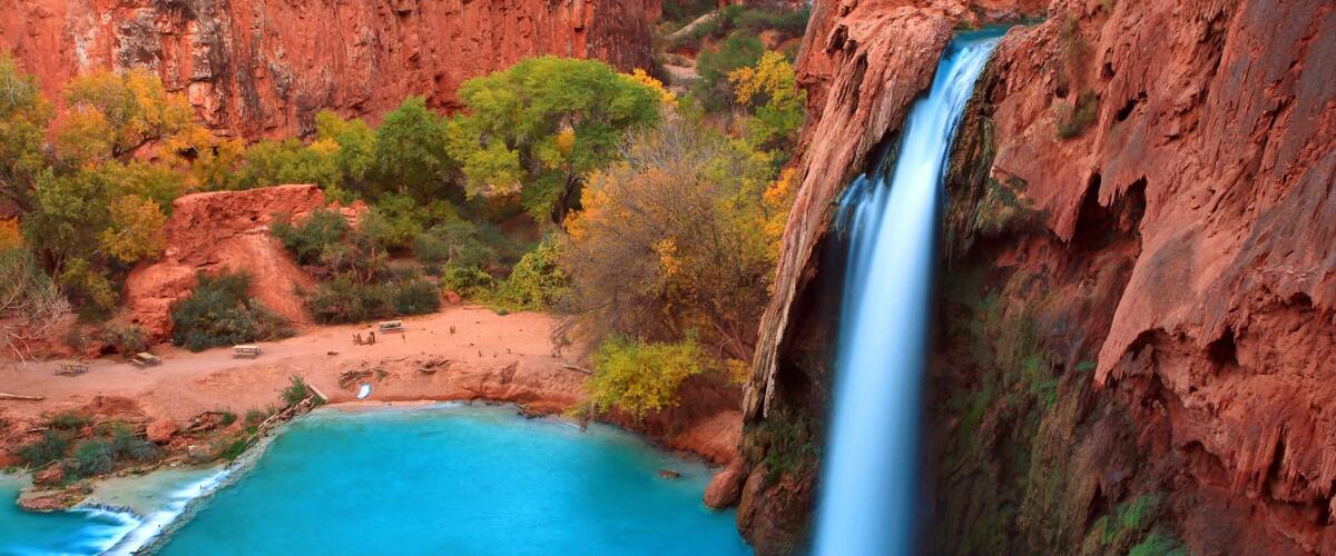 A waterfall cascading into a turquoise pool surrounded by red rock cliffs and autumn-colored trees.