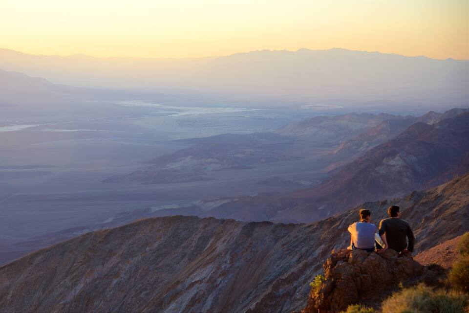 Two people sitting on a mountain ridge overlooking a valley with a river and distant mountains.