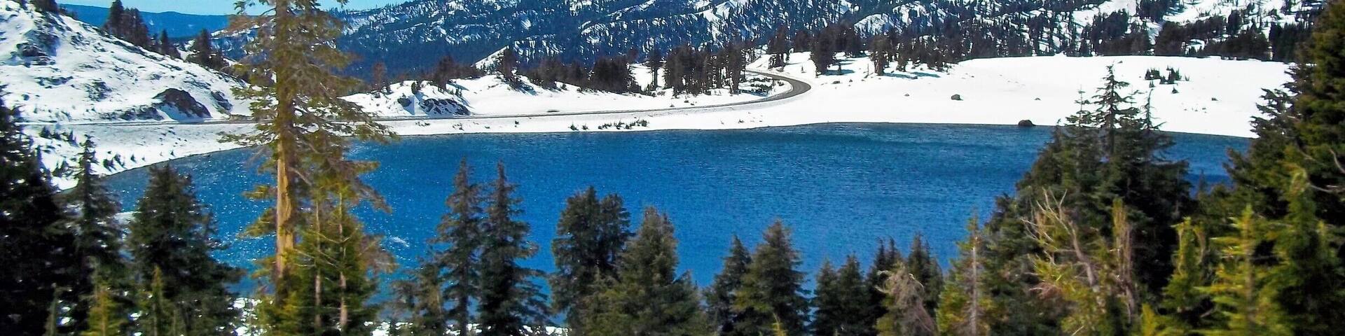 A snow-covered mountain range with a lake and dense forest in the foreground.