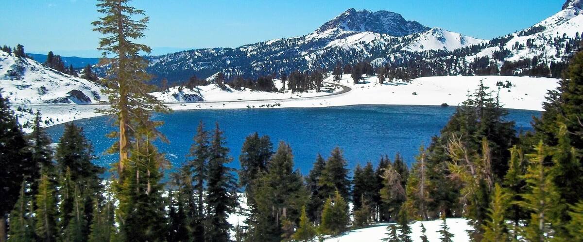 A snow-covered mountain range with a lake and dense forest in the foreground.