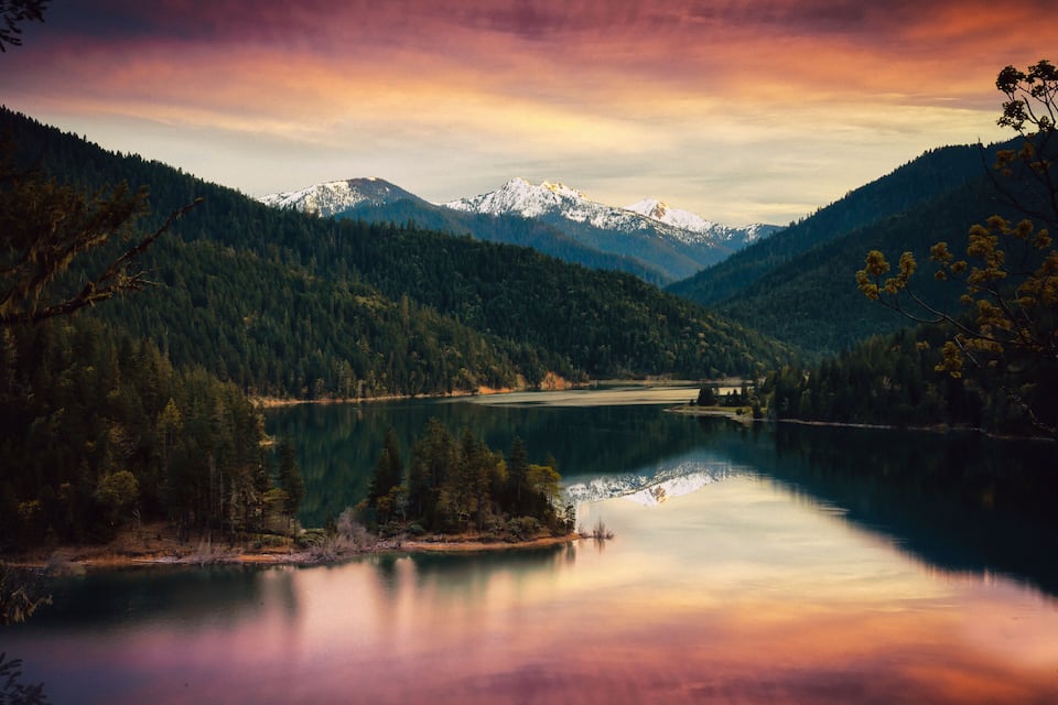 A serene lake surrounded by dense forests and snow-capped mountains under a colorful sky.