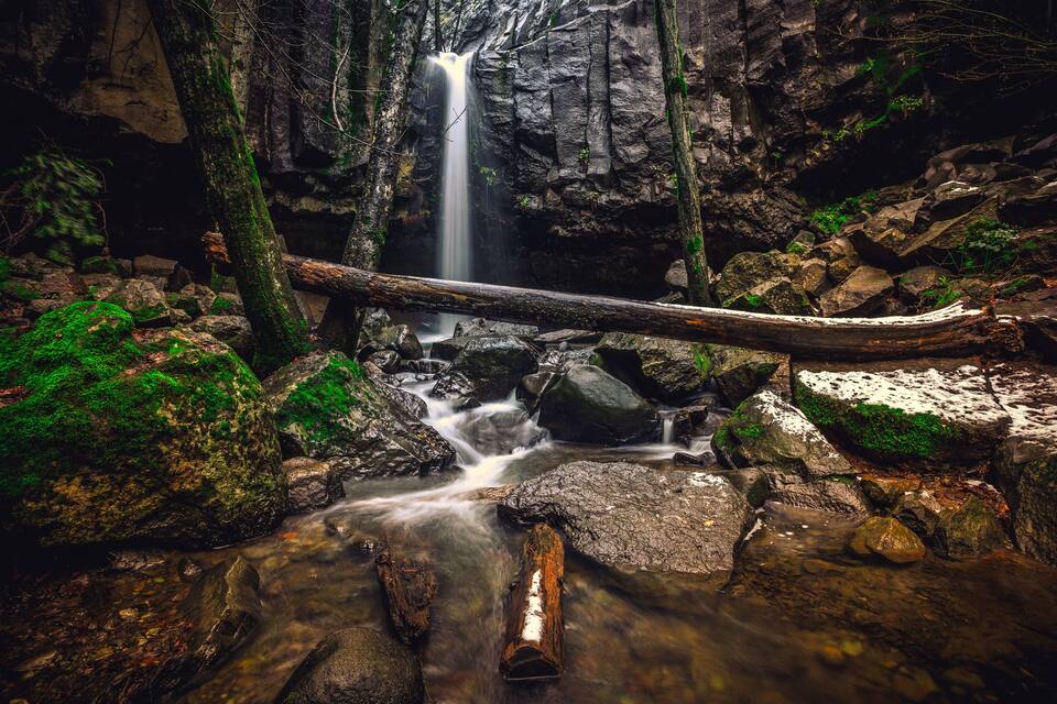 A waterfall in a forested area with moss-covered rocks and a fallen log.