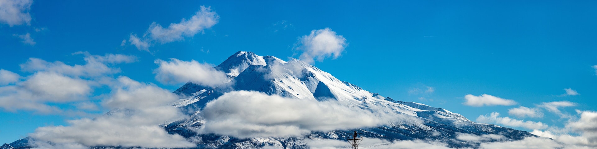 Pico de montanha coberto de neve, com nuvens ao redor da base e uma torre de transmissão em primeiro plano.