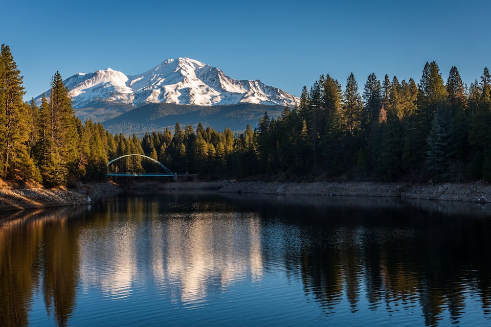 Um lago tranquilo com uma ponte, montanhas cobertas de neve e uma floresta densa ao fundo.