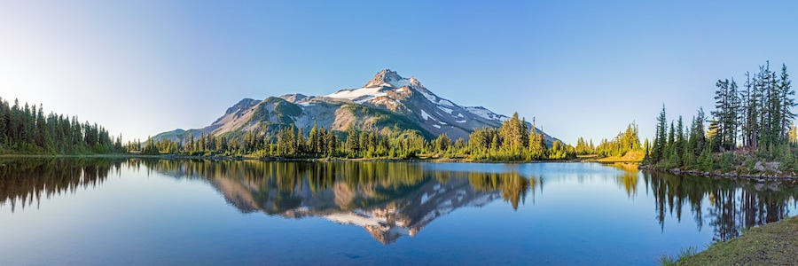 A mountain lake with clear reflections, surrounded by dense forest and snow-capped peaks.