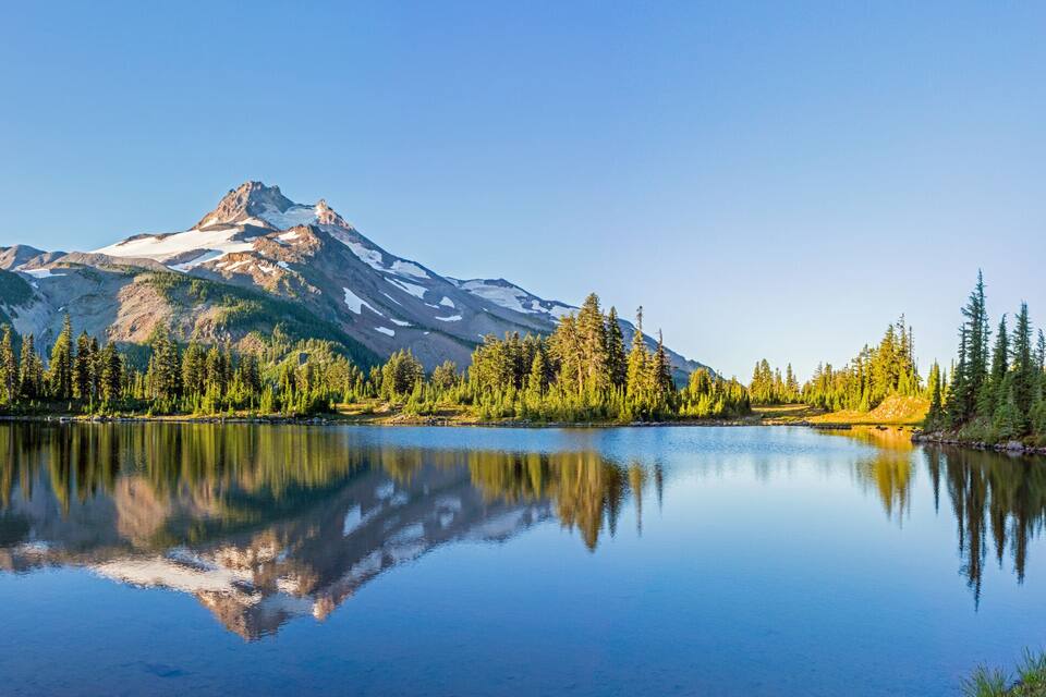 A mountain lake with clear reflections, surrounded by dense forest and snow-capped peaks.