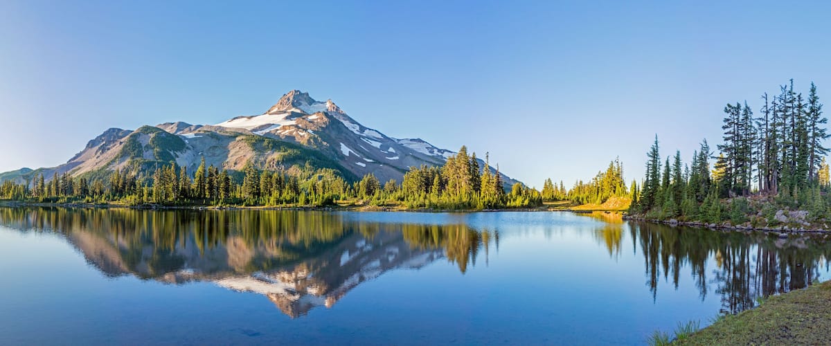 A mountain lake with clear reflections, surrounded by dense forest and snow-capped peaks.