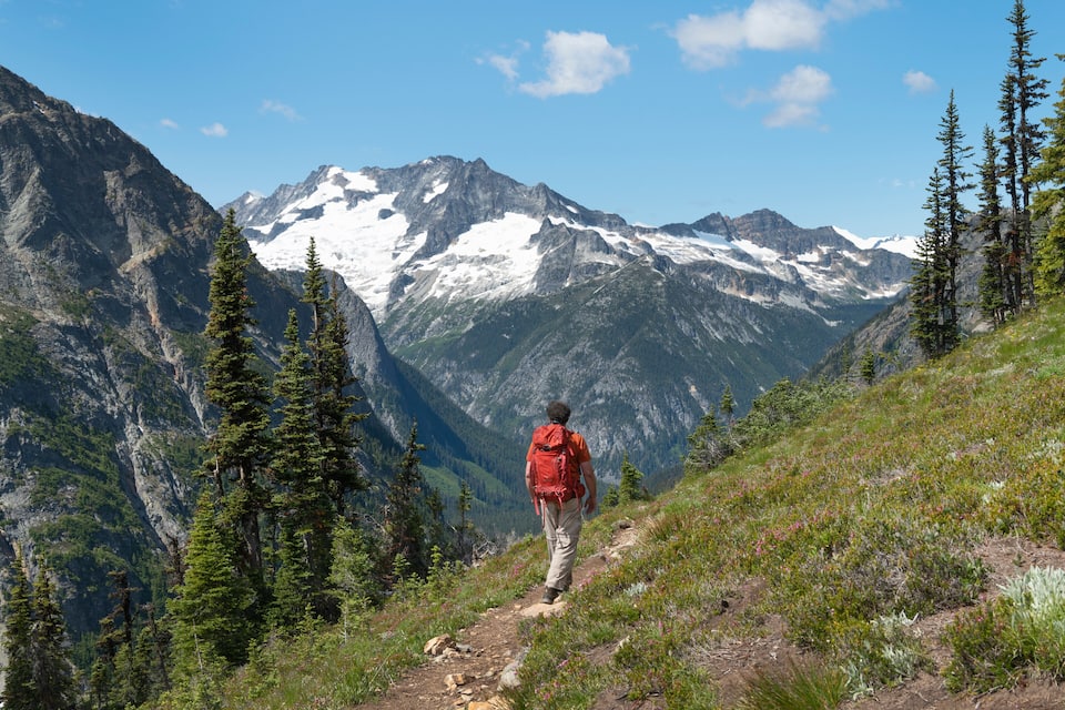 A hiker on a mountain trail with snow-capped peaks in the background.