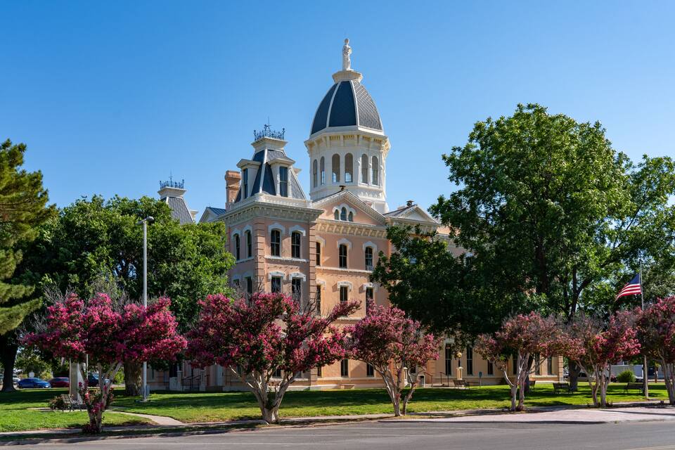 A historic building with a large dome and multiple towers, surrounded by lush green trees and a clear blue sky.