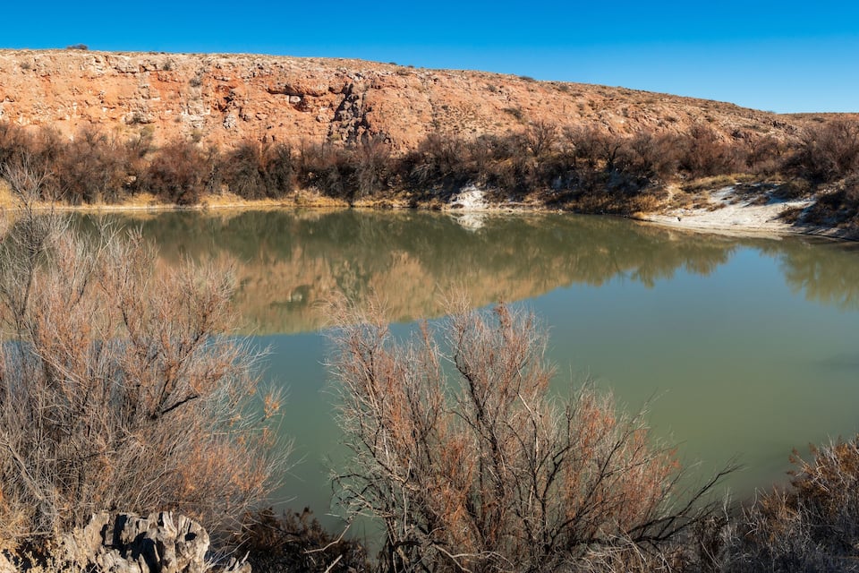 A calm lake surrounded by dry, sparse vegetation and a rocky hill.