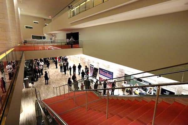 Espaço público interno com vários níveis, escada com carpete vermelho e guarda-corpos de vidro, com várias pessoas.