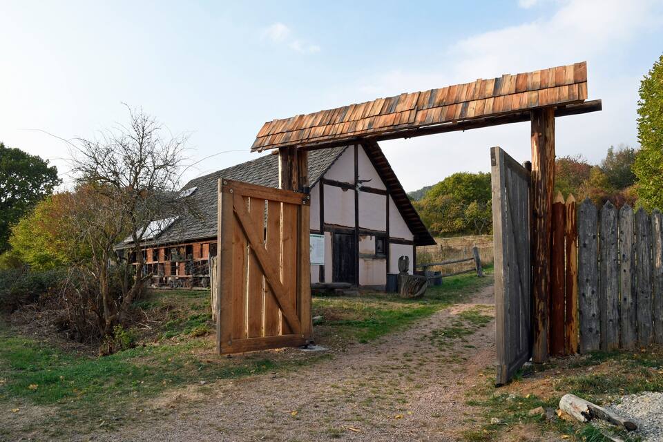 A rustic wooden structure with a slanted roof, a wooden gate, and a building with a gabled roof in the background.
