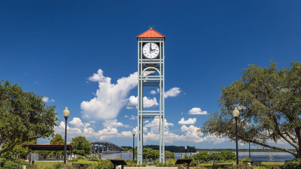 A clock tower with a red roof, surrounded by benches and a park with trees and a lake in the background.