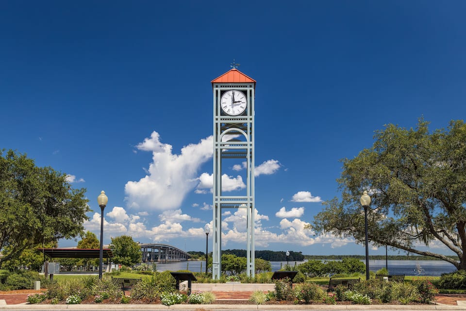 A clock tower with a red roof, surrounded by benches and a park with trees and a lake in the background.