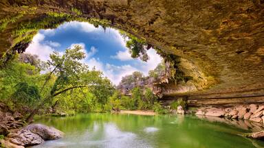 Um rio calmo sob um arco natural, rodeado por vegetação exuberante e terreno rochoso.