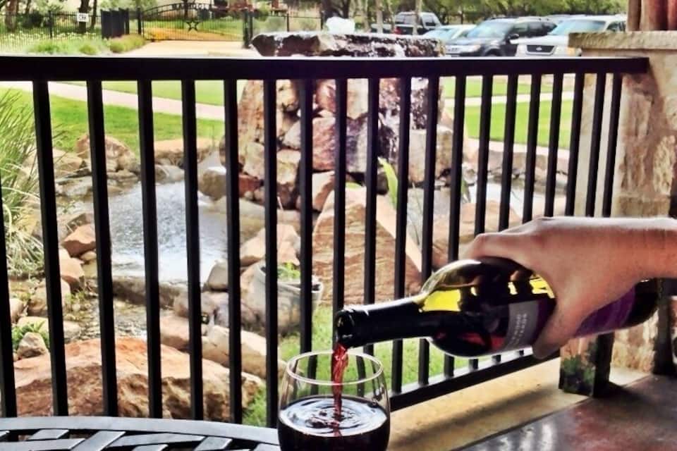 A hand pouring red wine into a glass on a metal table with a view of a rocky stream and trees in the background.
