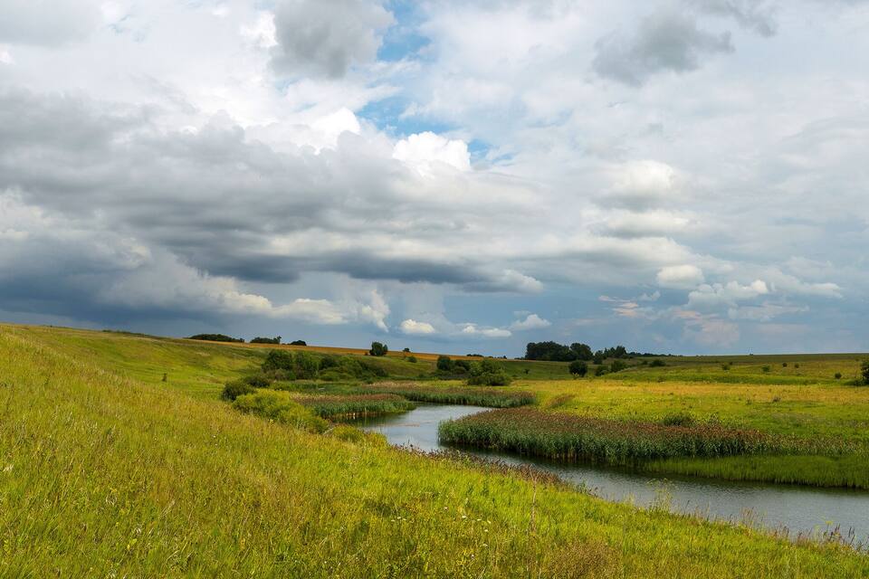 Ein Fluss schlängelt sich durch ein üppiges Grünfeld, über dem sich eine bewölkte Himmeldecke erstreckt.